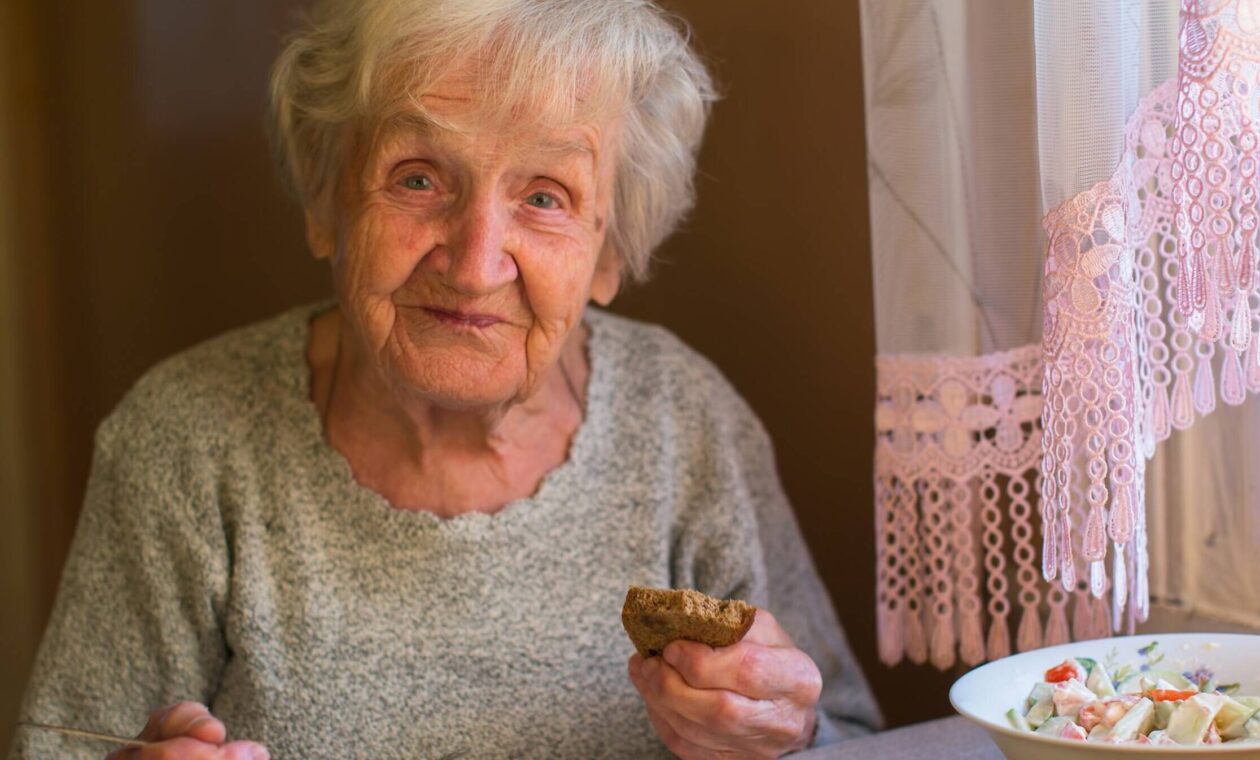 Äldre dam med vitt hår sitter och äter vid sitt köksbord.