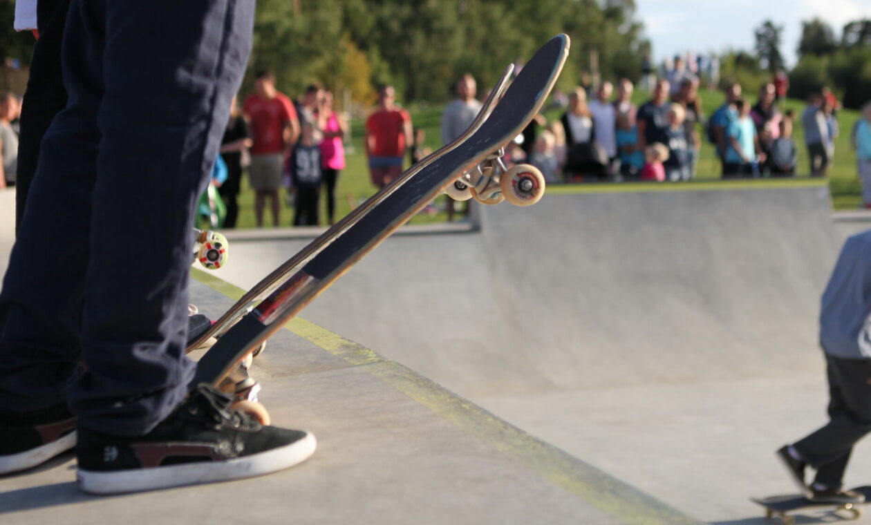 Närbild på ben i jeans och gympaskor med skateboard vid ramp i betong. Publik i förgrunden och kille på skateboard.