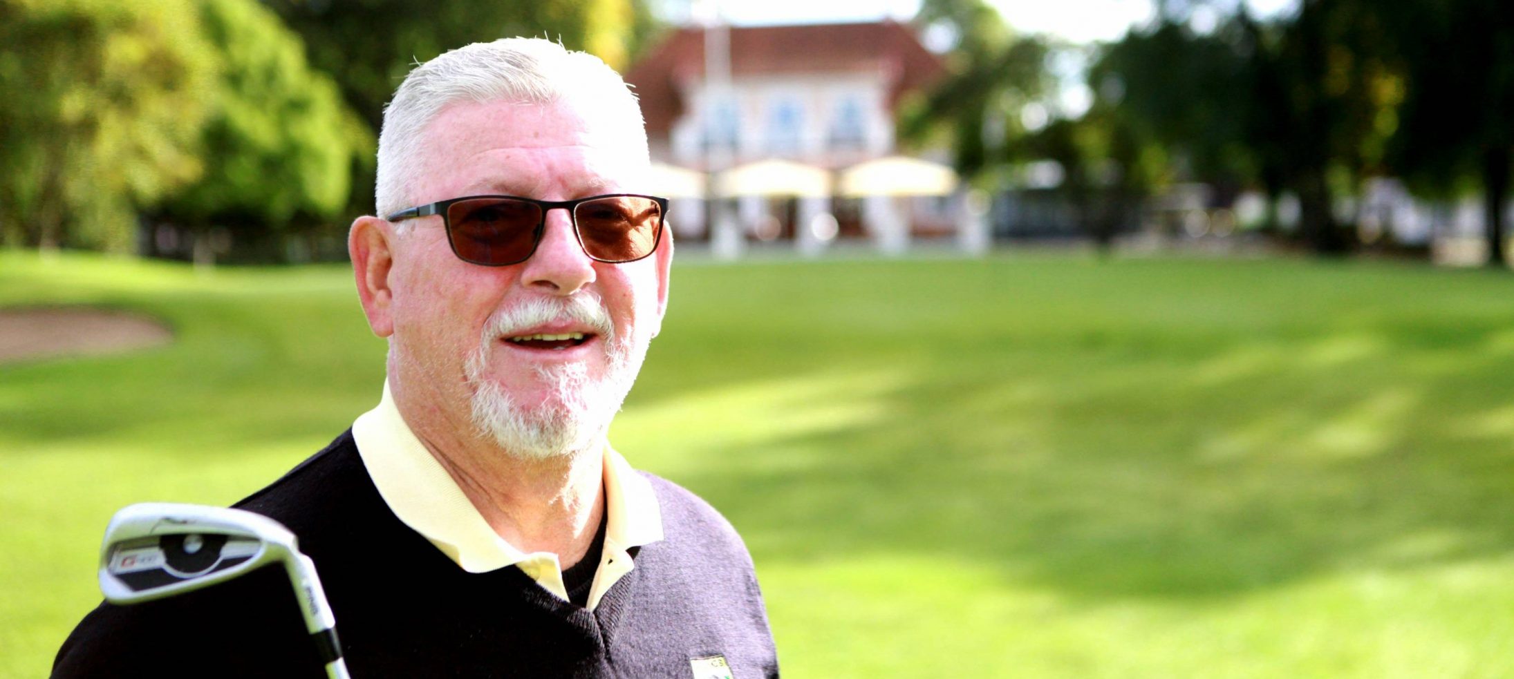 Senior med golfklubba, Leif Ziegler, med solglasögon framför golfbana och klubbhus på Kävlinge GK