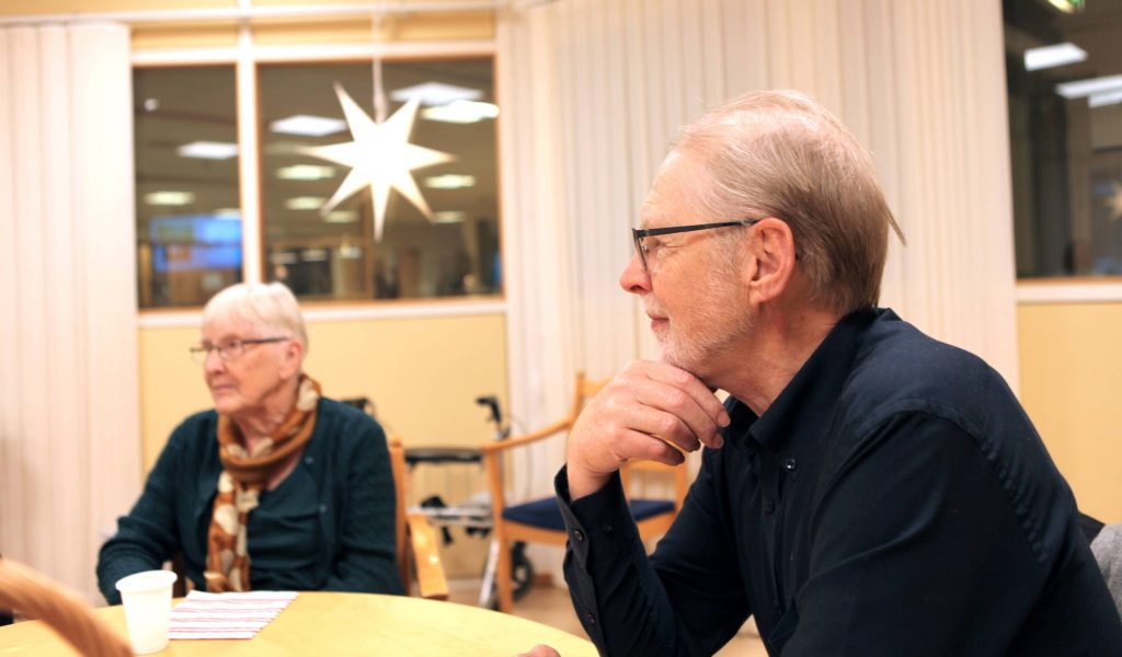Äldre man med skägg och glasögon i profil samt äldre kvinna i förgrunden sittandes vid ett konferensbord.